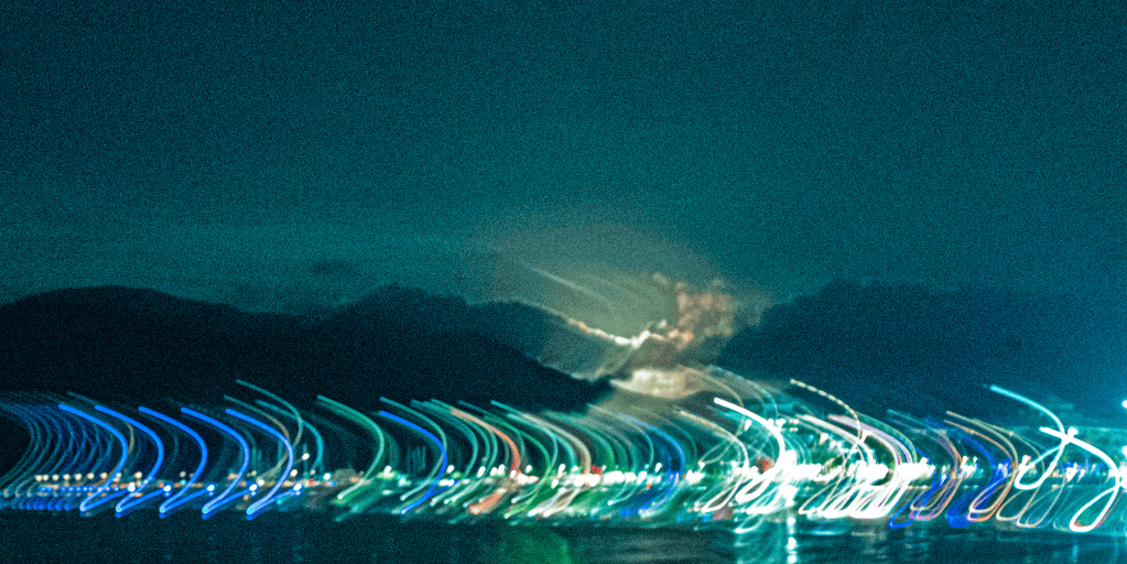 Celestial Vespers Evening Song | DANGEROUS BEAUTY Long-exposure night cityscape with colorful curved light trails along the water and dark mountains in the distance.